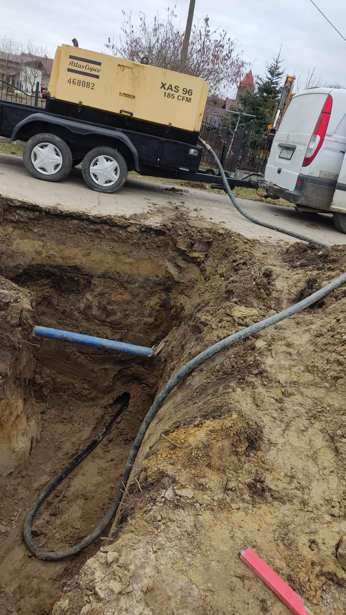 Głęboki wykop w ziemi z widocznymi rurami: niebieską i czarną, z żółtym kompresorem Atlas Copco XAS 96 185 CFM na przyczepie w tle i częściowo widocznym białym vanem.