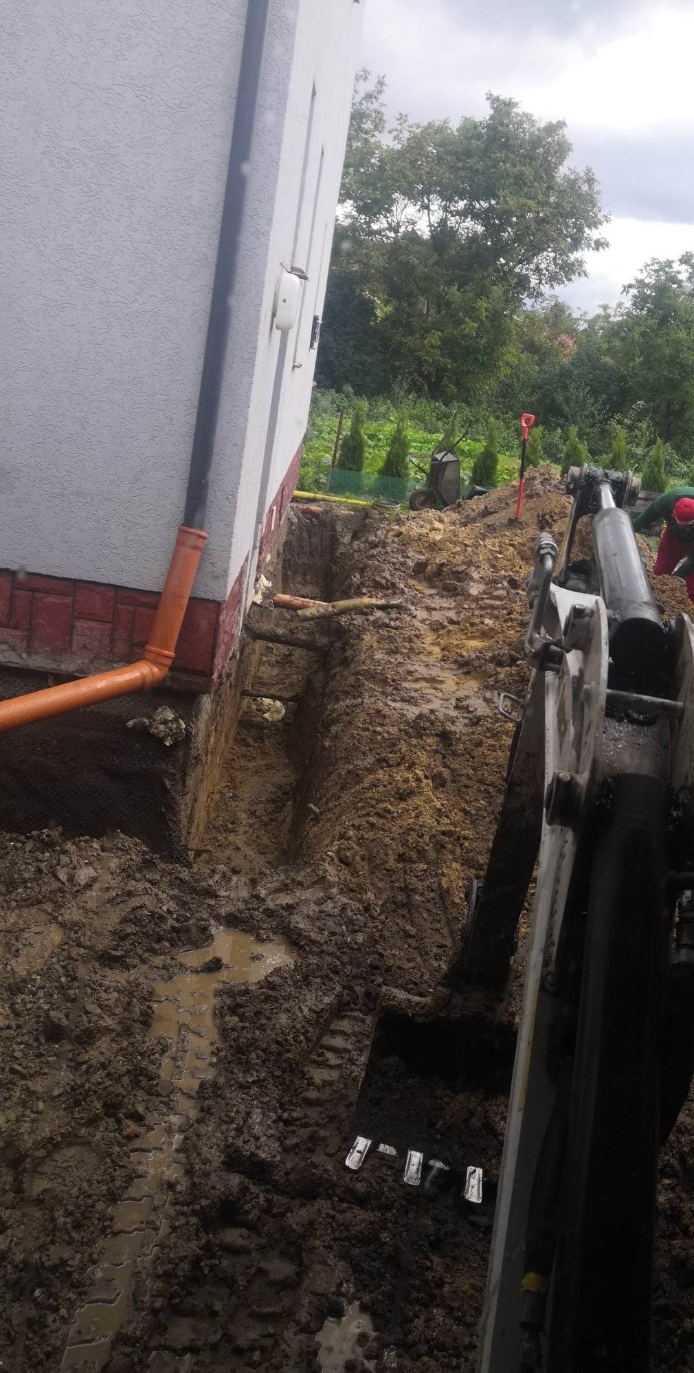 Wykop liniowy wzdłuż fundamentu budynku, widoczna pomarańczowa rura drenażowa, fragment koparki i błotniste podłoże.