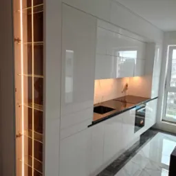 Nowoczesna, biała kuchnia z połyskiem, podświetlane szklane półki w szafce, czarny blat i marmurowy backsplash, widok z góry na podłogę z płytkami imitującymi marmur.