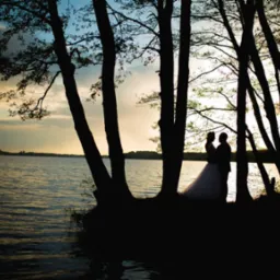 Siluety nowożeńców na brzegu jeziora o zachodzie słońca, widoczne przez pnie drzew.