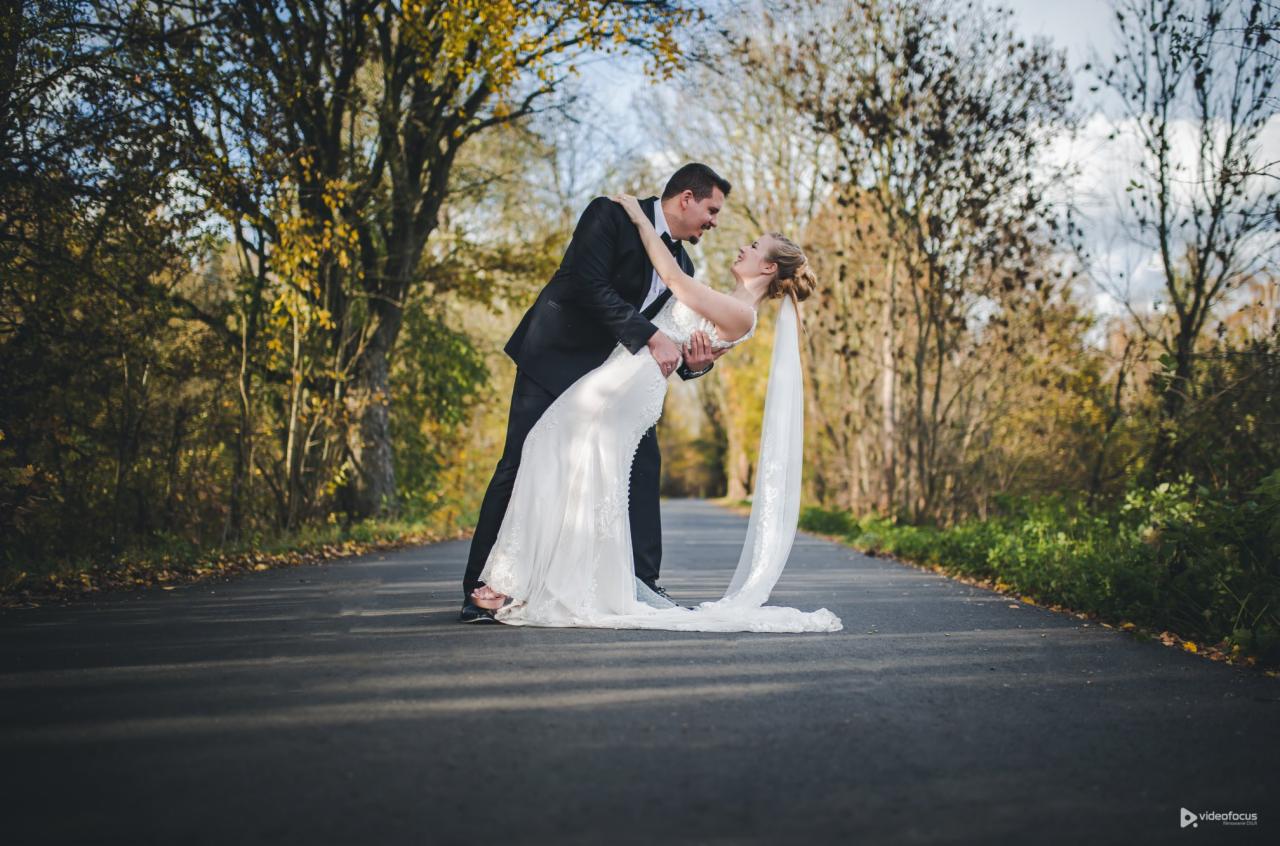 Para młoda w tańcu na asfaltowej drodze otoczonej drzewami w jesiennej scenerii, panna młoda w białej sukni z długim welonem, pan młody w czarnym garniturze.