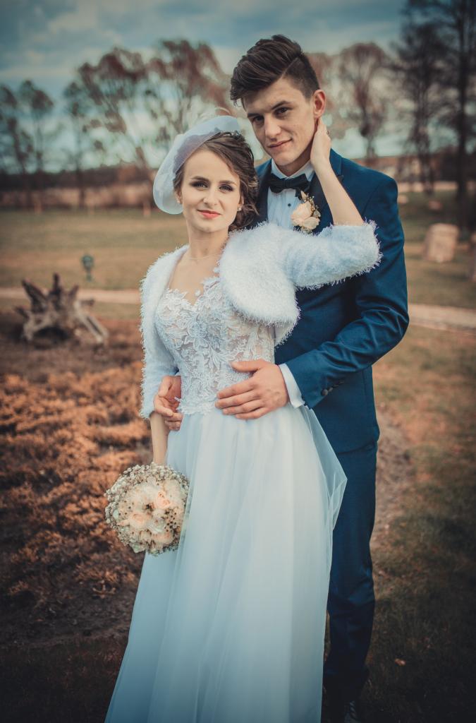 Para młoda: Panna młoda w koronkowej sukni z futrzanym bolerkiem i welonem, trzyma bukiet. Pan młody w granatowym garniturze z butonierką, obejmuje ją. Tło: park z drzewami.