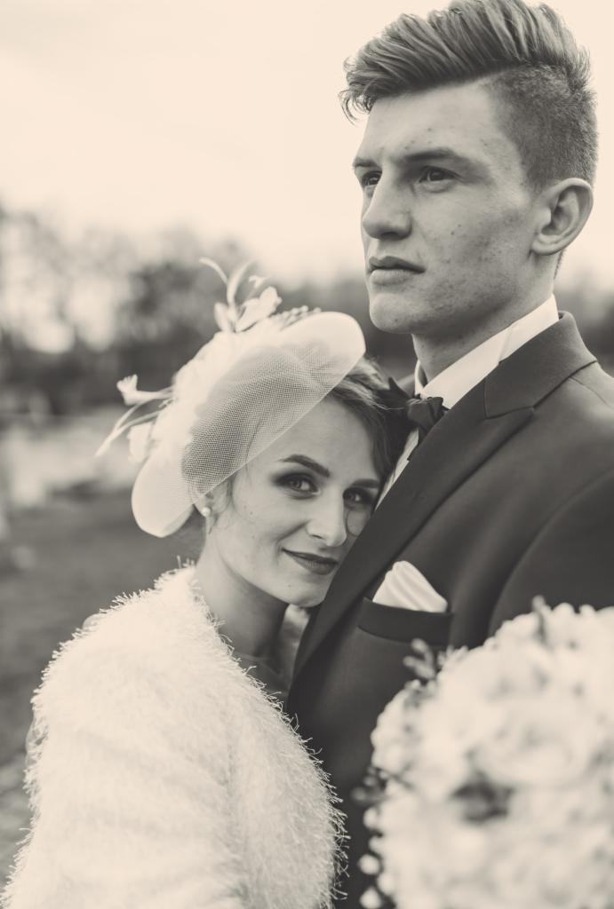 Czarno-białe zdjęcie pary młodej: panna młoda w kapeluszu z woalką i futrzanym okryciem, opiera głowę o ramię pana młodego w ciemnym garniturze z butonierką i białą poszetką; bukiet ślubny w kadrze.