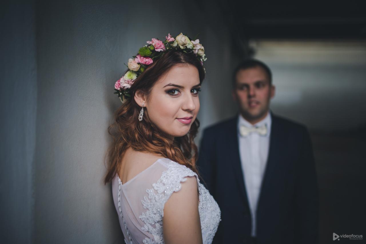 Panna młoda w wianku z kwiatów i koronkową suknią, spogląda przez ramię, w tle rozmyty pan młody w ciemnym garniturze i muszce.