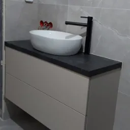 Nowoczesna łazienka w szarościach: biała umywalka nablatowa na szarym blacie z szafką, czarna bateria, szare płytki imitujące marmur, widoczne niedokończone prace instalacyjne.