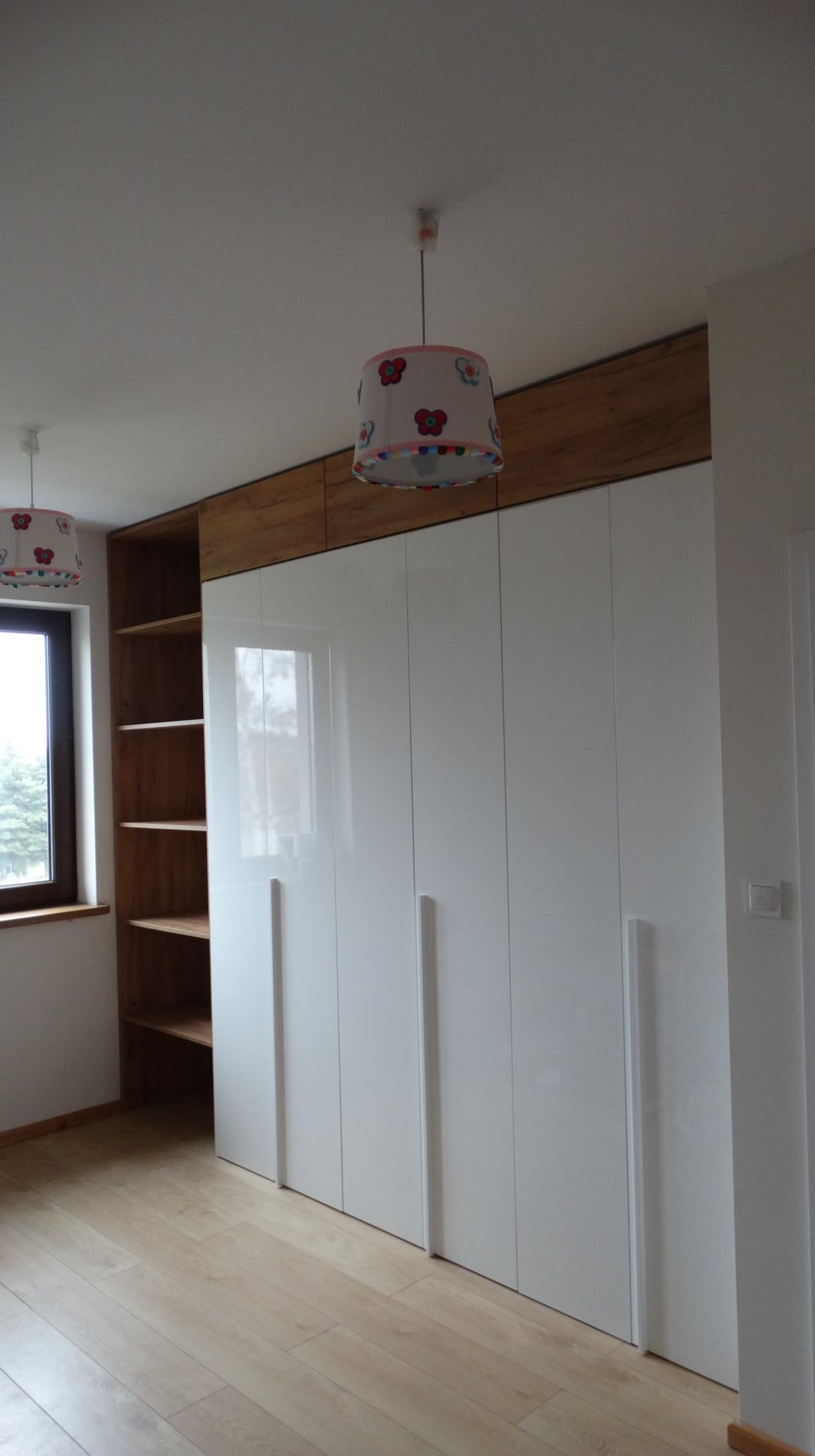 Biała szafa wnękowa z połyskiem, z drewnianym wykończeniem u góry, w pokoju z drewnianą podłogą i lampami z motywem kwiatowym.