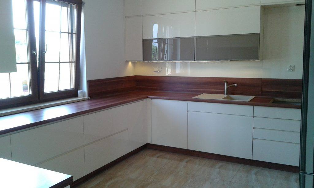 Nowoczesna kuchnia z białymi, lakierowanymi frontami szafek i drewnianym blatem, oświetlenie LED pod szafkami wiszącymi i okno z drewnianą ramą.