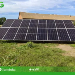 Panel słoneczny o mocy 9kW na trawiastym terenie, z widocznym budynkiem gospodarczym w tle i niebieskim niebem.