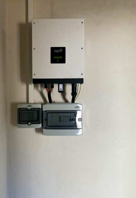 Biały inwerter marki FOX z czarnym wyświetlaczem, zamontowany na ścianie nad dwiema szarymi skrzynkami elektrycznymi z widocznymi przewodami.