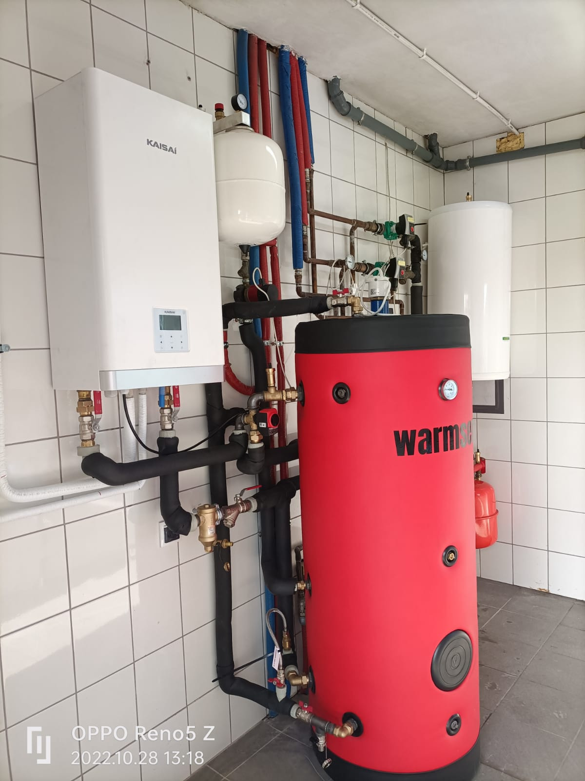 Kompletna instalacja grzewcza w pomieszczeniu z białymi kafelkami, widoczny biały piec Kaisai, czerwony zbiornik akumulacyjny Warmer i biały zbiornik wyrównawczy. Rury miedziane i czarne izolowane...