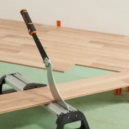 Przycinanie panela podłogowego za pomocą specjalistycznego noża na tle zielonej pianki izolacyjnej i ułożonej części podłogi z dystansami przy ścianie.