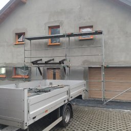 Pyzik Krzysztof - Ściana domu w trakcie tynkowania, z oknami zabezpieczonymi folią i rusztowaniem. Przyczepa z narzędziami na pierwszym planie. Widoczne ślady prac budowlanych.