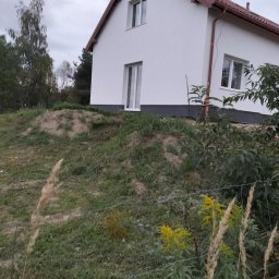 Pyzik Krzysztof - Biały dom jednorodzinny z brązowym dachem, widziany z dołu, na tle pochmurnego nieba i zieleni. Widoczne okna i rynny. Dom po remoncie elewacji.