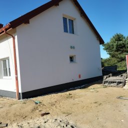 Pyzik Krzysztof - Boczny widok otynkowanego, dwukondygnacyjnego domu z brązowym dachem i rynnami. Widoczne okna, fragment elewacji i teren wokół budynku z piaskiem i rusztowaniami.