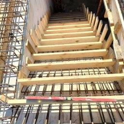 Pyzik Krzysztof - Zbrojenie fundamentów z drewnianym szalunkiem schodów, widok z góry. Czerwona poziomica na zbrojeniu. Elementy konstrukcyjne w surowym stanie.