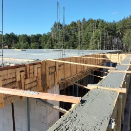 Pyzik Krzysztof - Wylany beton na szalunku fundamentów z widocznymi ścianami z bloczków i zbrojeniami. W tle las i błękitne niebo. Prace budowlane w toku, etap fundamentów.