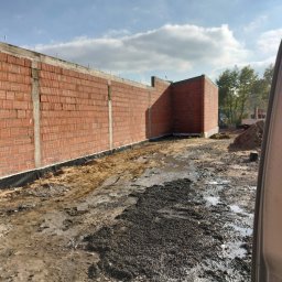 Wznoszenie muru z czerwonej cegły z widocznymi spoinami i izolacją przeciwwilgociową u podstawy, na tle nieba z chmurami i fragmentem pojazdu z prawej strony.