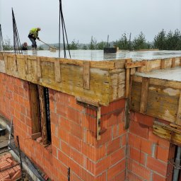 Murowanie ścian z czerwonej cegły, zalewanie betonem stropu z drewnianym szalunkiem. Dwóch pracowników w trakcie pracy na budowie w pochmurny dzień.