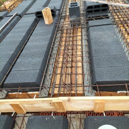 Budowa stropu: szare bloczki, zbrojenie z prętów, drewniane elementy szalunku. Widoczny poziomica i wiadro ze śrubami. Ujęcie z góry, w trakcie prac.