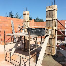 Pyzik Krzysztof - Szare rusztowanie, drewniane szalunki kolumn z wystającymi prętami zbrojeniowymi, czarna kuweta na zaprawę, mur z czerwonej cegły dziurawki w tle, błękitne niebo.