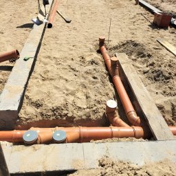 Pyzik Krzysztof - Instalacja kanalizacyjna w piasku, widoczne rury z szarymi pokrywami rewizyjnymi, częściowo zakopane w wykopie fundamentowym. Widok z góry, słoneczny dzień.