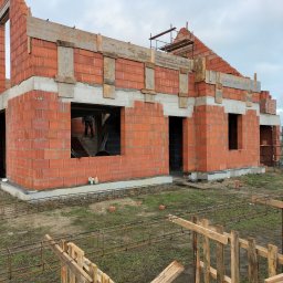 Pyzik Krzysztof - Surowy stan domu z czerwonej cegły, widoczne zbrojenia fundamentów i drewniane szalunki. W tle pochmurne niebo i częściowo widoczna osoba wewnątrz budynku.