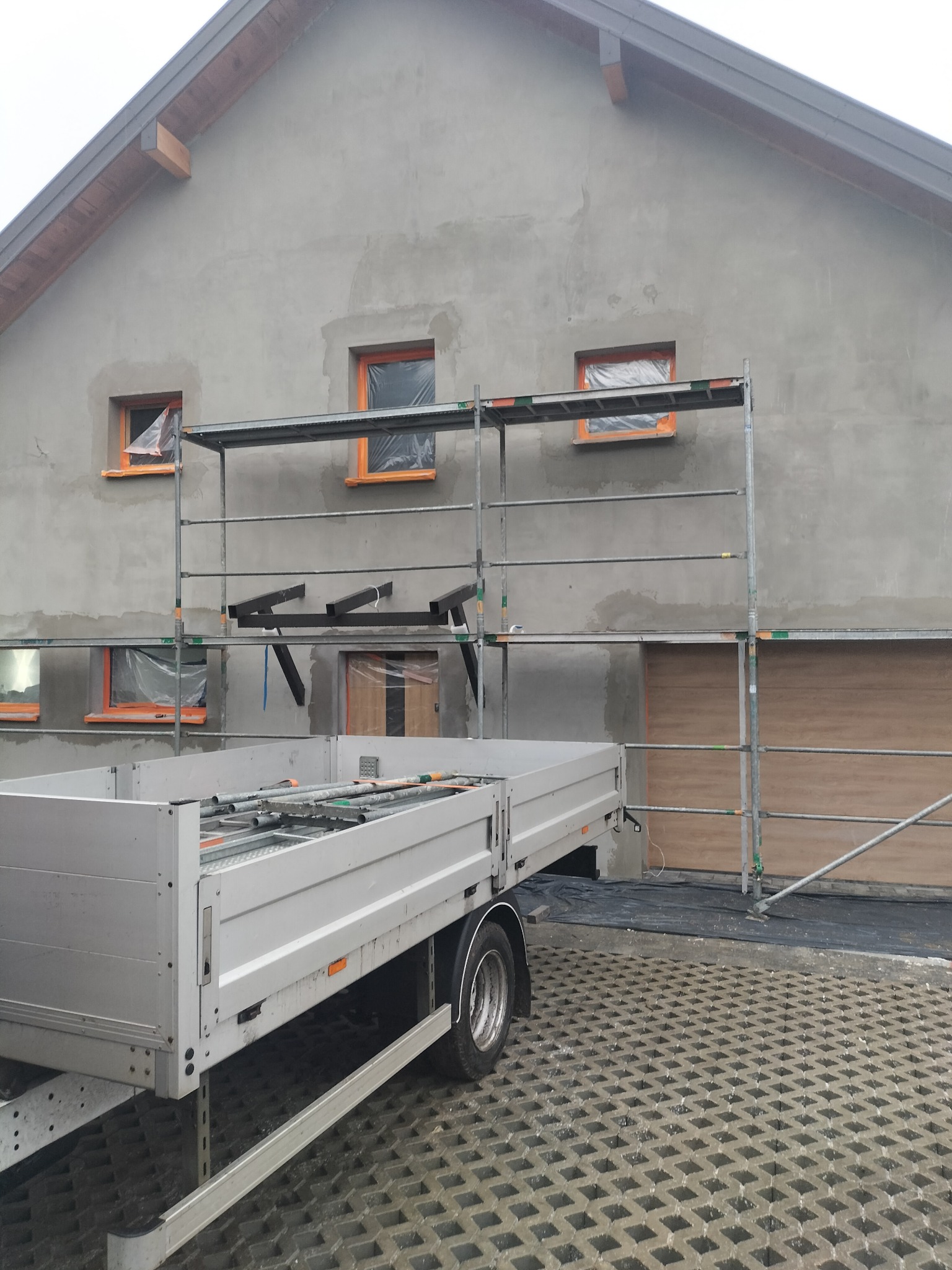 Ściana domu w trakcie tynkowania, z oknami zabezpieczonymi folią i rusztowaniem. Przyczepa z narzędziami na pierwszym planie. Widoczne ślady prac budowlanych.