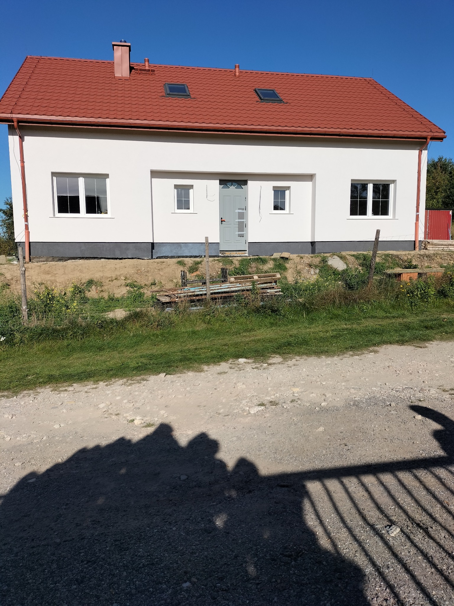 Biały dom z czerwoną dachówką i szarymi drzwiami wejściowymi w słoneczny dzień. Widoczne okna, komin i fragment terenu przed budynkiem.