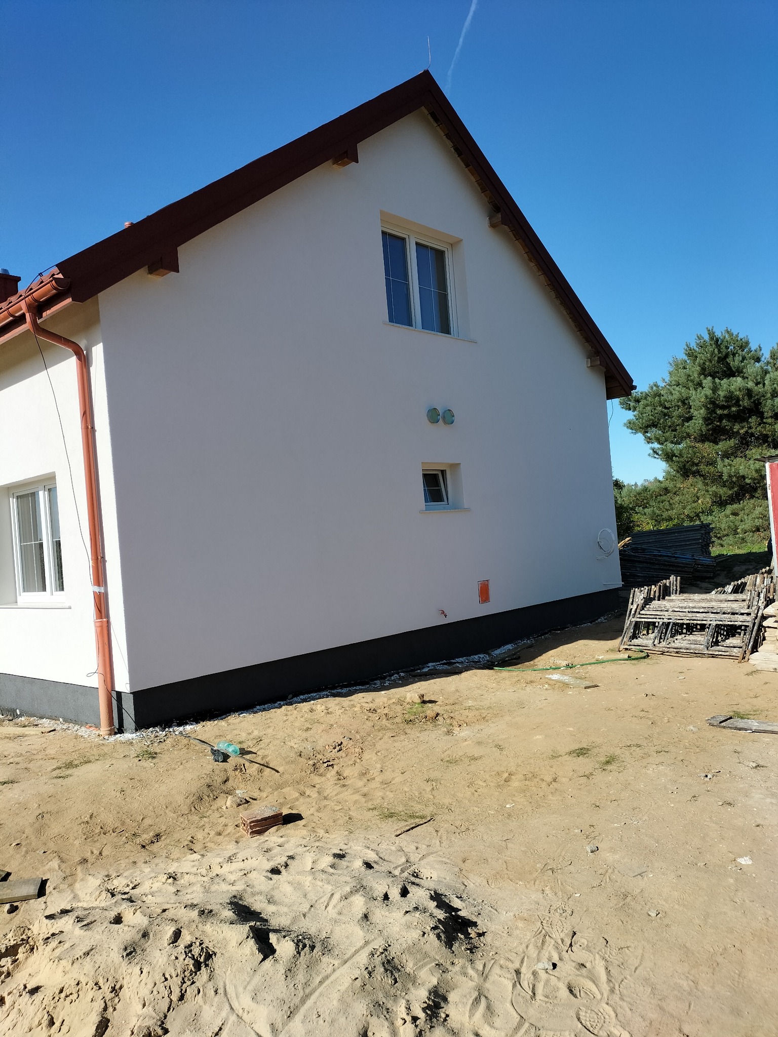 Boczny widok otynkowanego, dwukondygnacyjnego domu z brązowym dachem i rynnami. Widoczne okna, fragment elewacji i teren wokół budynku z piaskiem i rusztowaniami.