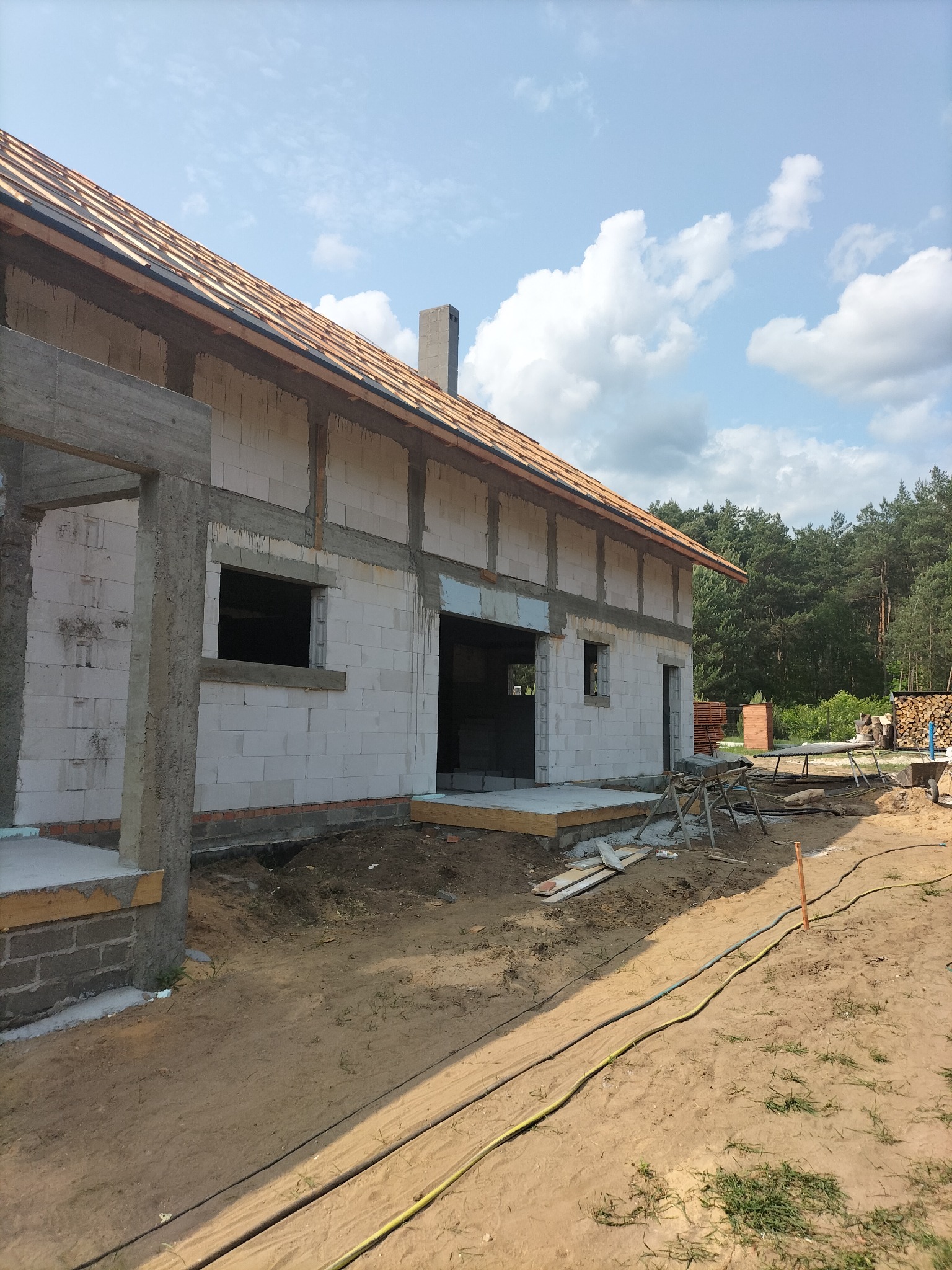 Budowa domu jednorodzinnego w stanie surowym otwartym. Widoczne ściany z bloczków, drewniana konstrukcja dachu i fundamenty. W tle las i błękitne niebo z chmurami.