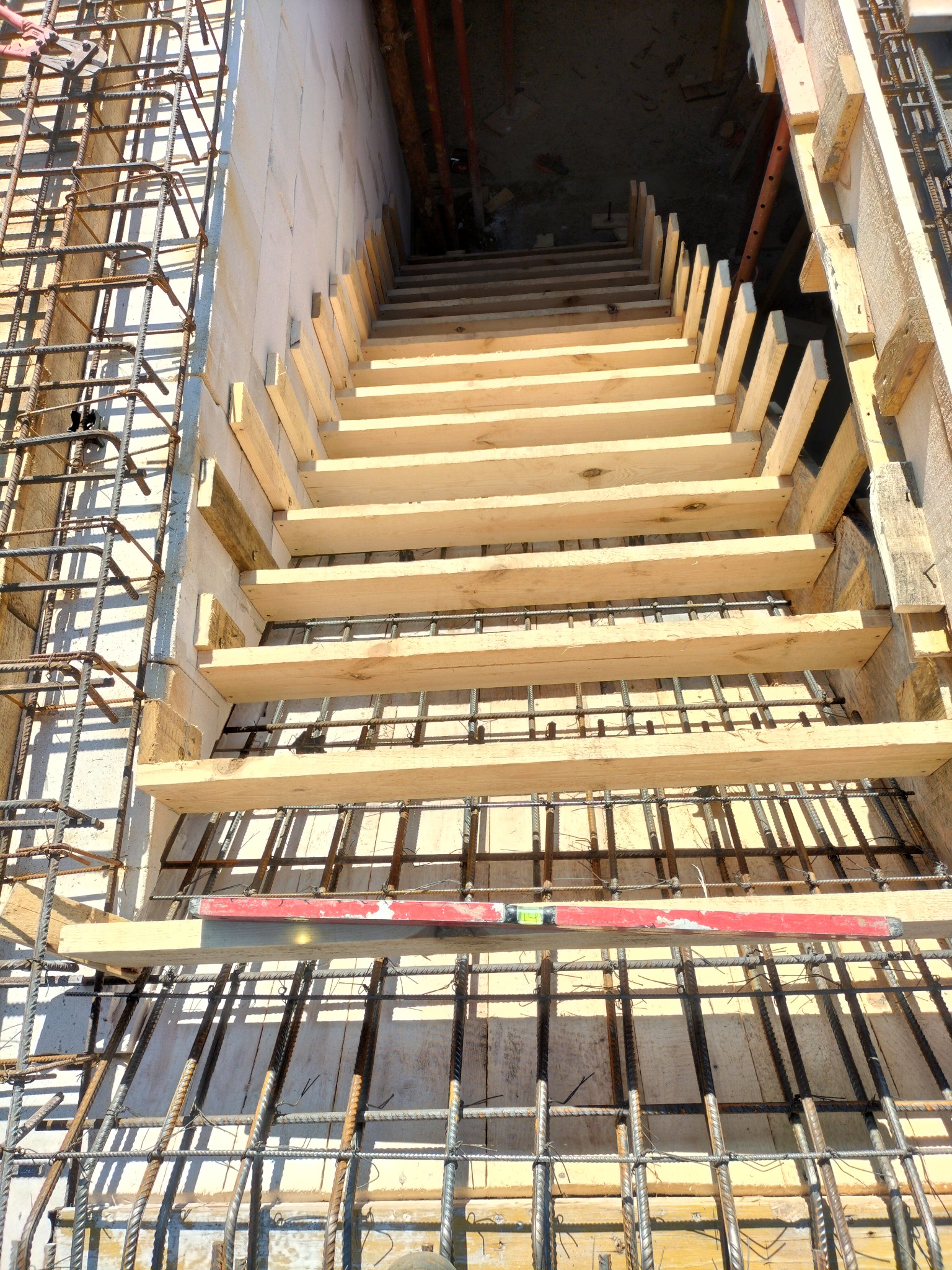Zbrojenie fundamentów z drewnianym szalunkiem schodów, widok z góry. Czerwona poziomica na zbrojeniu. Elementy konstrukcyjne w surowym stanie.