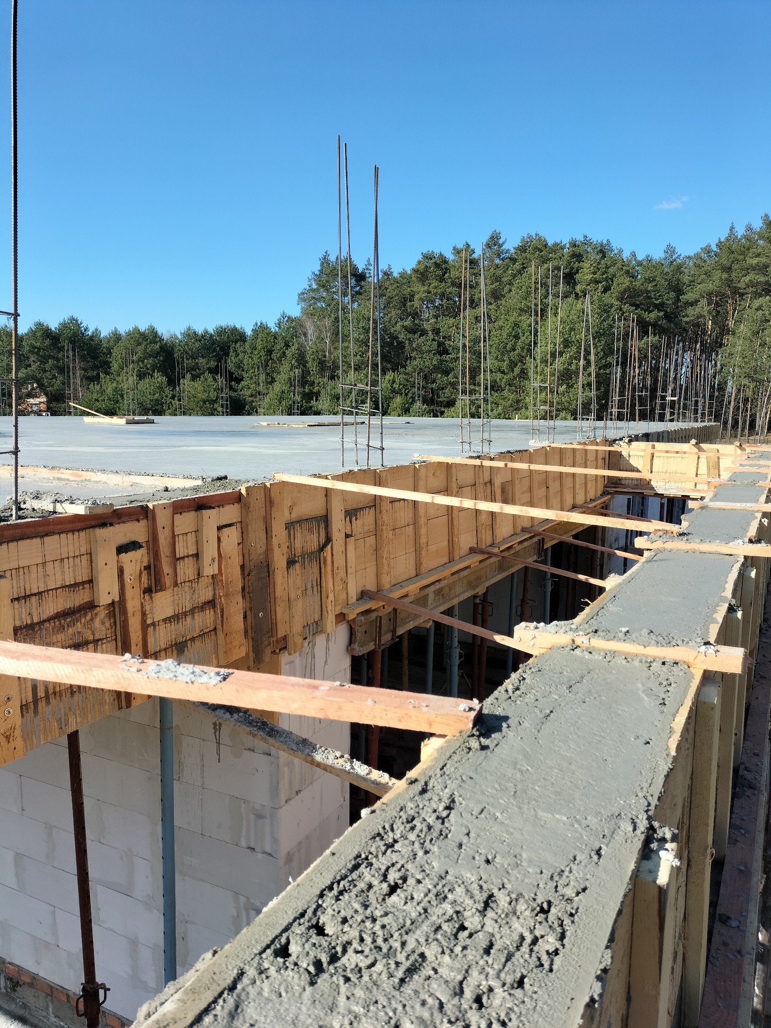 Wylany beton na szalunku fundamentów z widocznymi ścianami z bloczków i zbrojeniami. W tle las i błękitne niebo. Prace budowlane w toku, etap fundamentów.