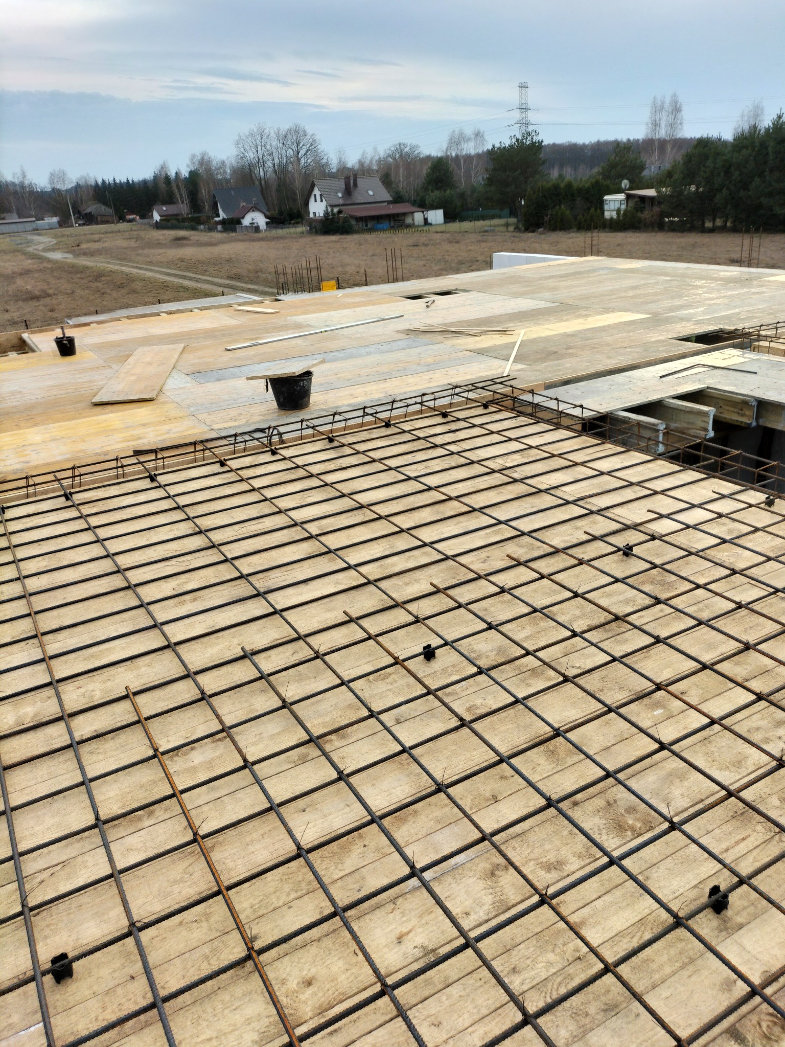 Zbrojenie płyty fundamentowej na deskowaniu z widocznymi elementami dystansowymi. W tle zabudowa jednorodzinna i pochmurne niebo.