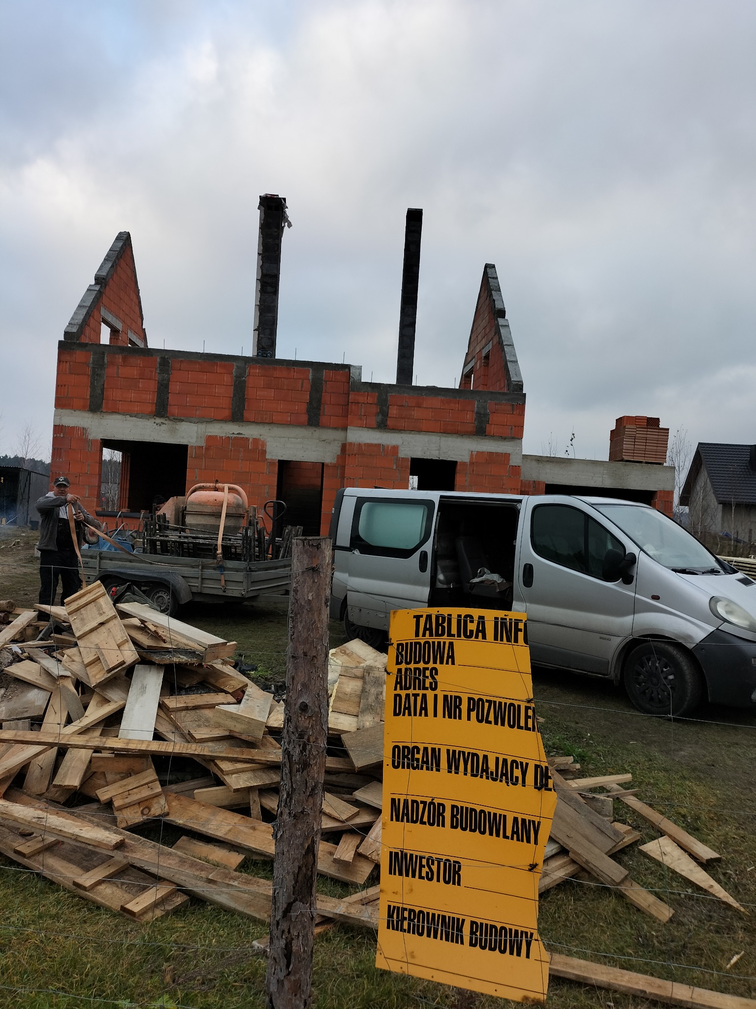 Budowa domu z czerwonej cegły w trakcie realizacji. Na pierwszym planie tablica informacyjna budowy, przyczepa z betoniarką i sterta drewna. Widoczny pracownik i srebrny van.