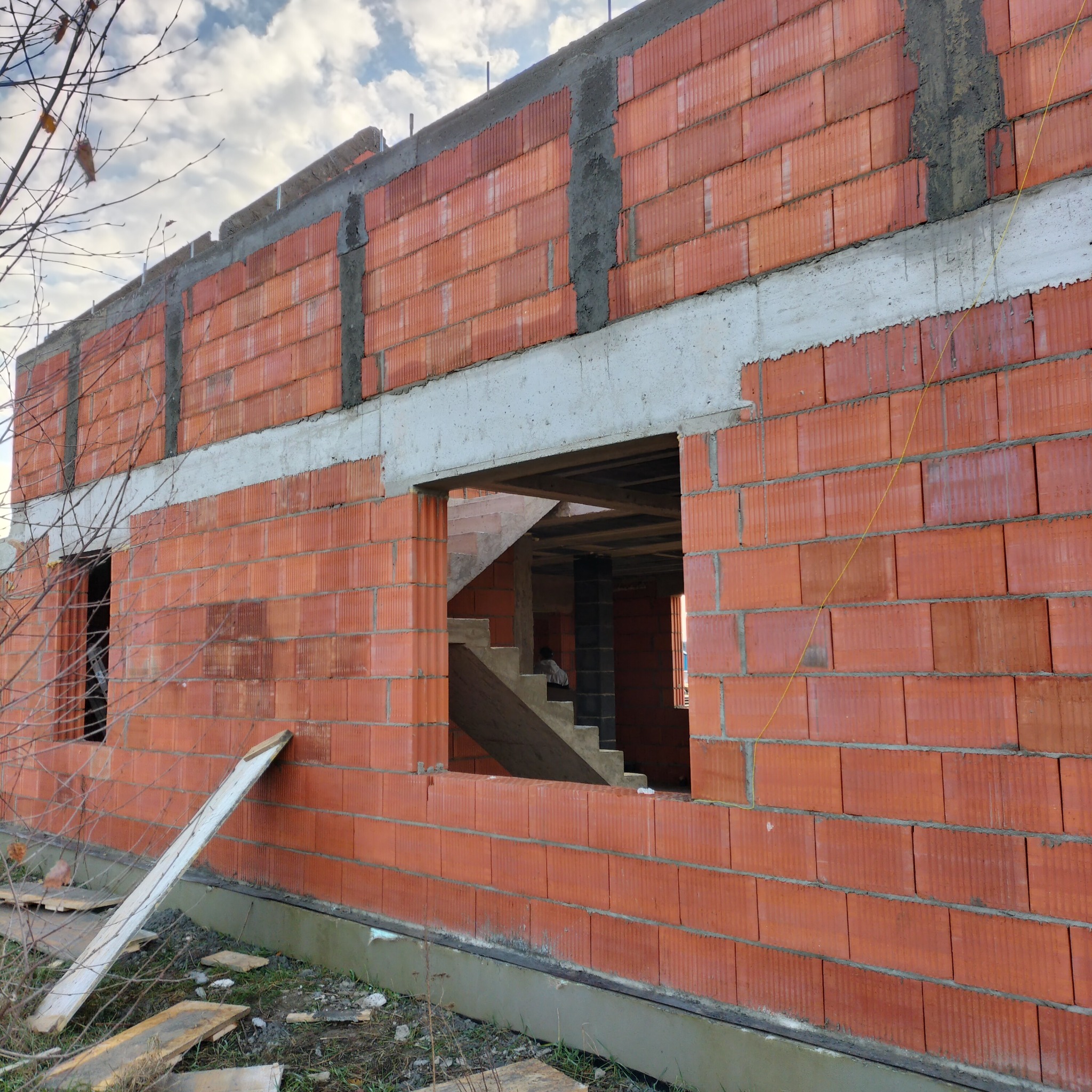 Wznoszenie ścian z czerwonej cegły ceramicznej porotherm, widoczne betonowe nadproża okienne i zbrojenia. W tle surowe schody wewnątrz budynku.