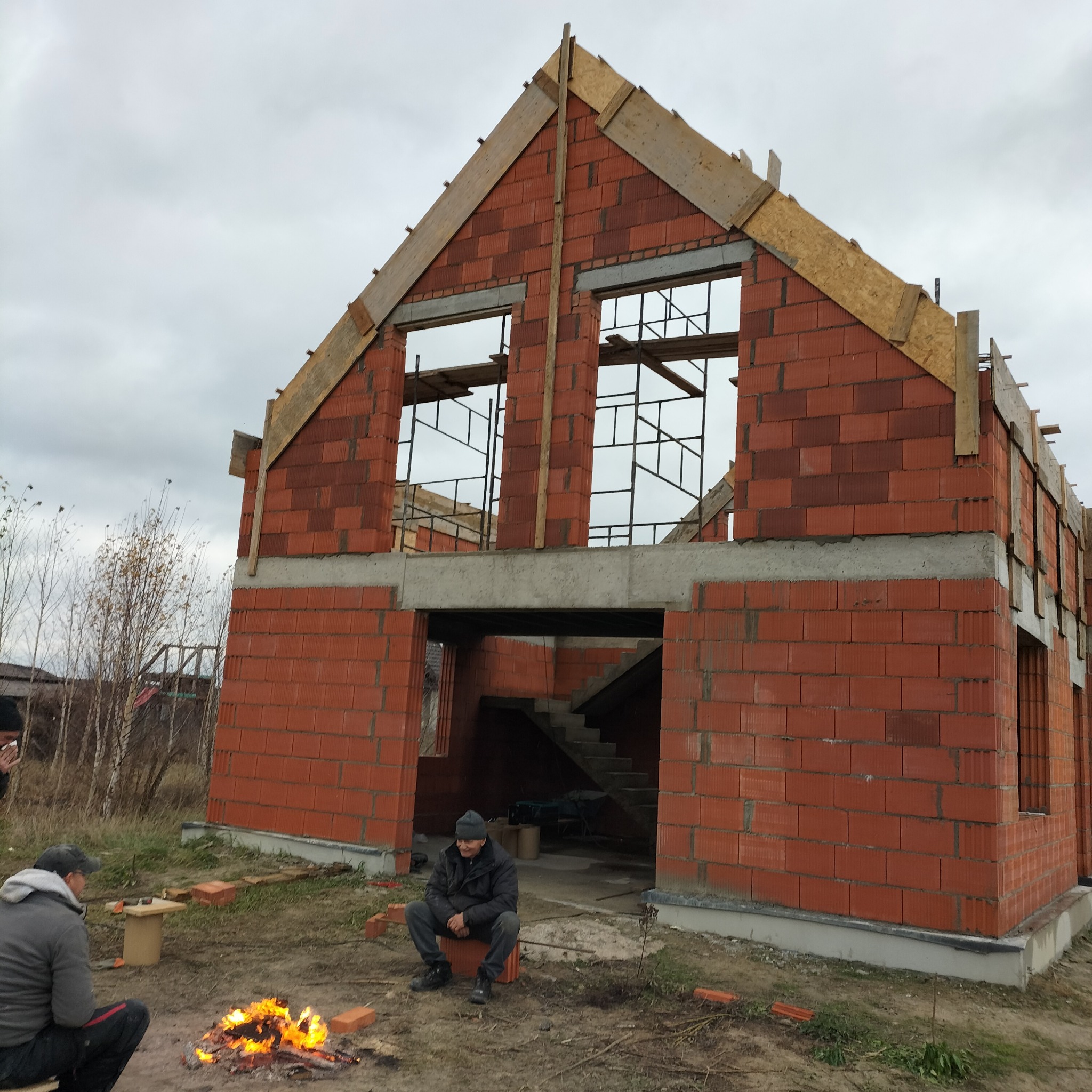 Budowa domu z czerwonej cegły w trakcie realizacji. Widoczne ściany, otwory okienne i drewniane elementy konstrukcyjne. Na pierwszym planie ognisko i odpoczywający robotnicy.