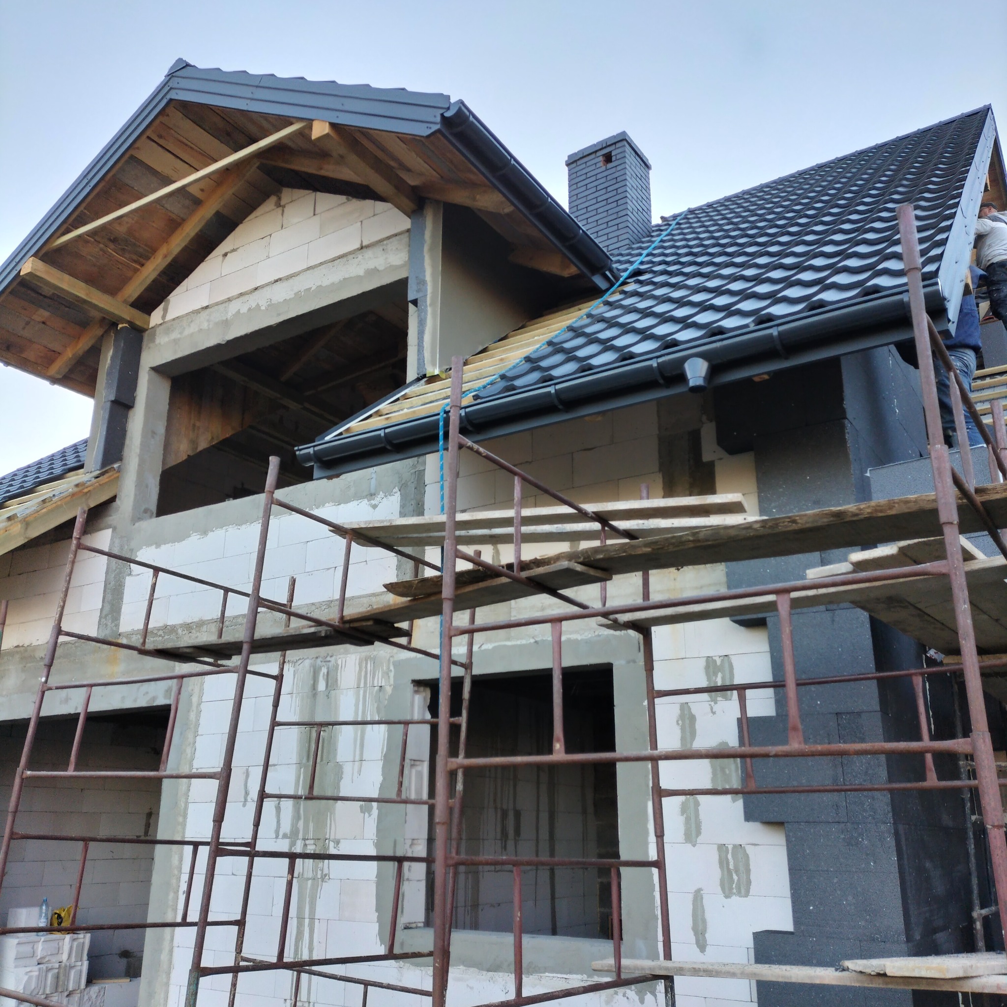 Budowa domu w trakcie: ściany z białych bloczków, częściowo ocieplone, rusztowania, dach z czarnej dachówki. Widoczny komin i fragment drewnianej konstrukcji dachu.