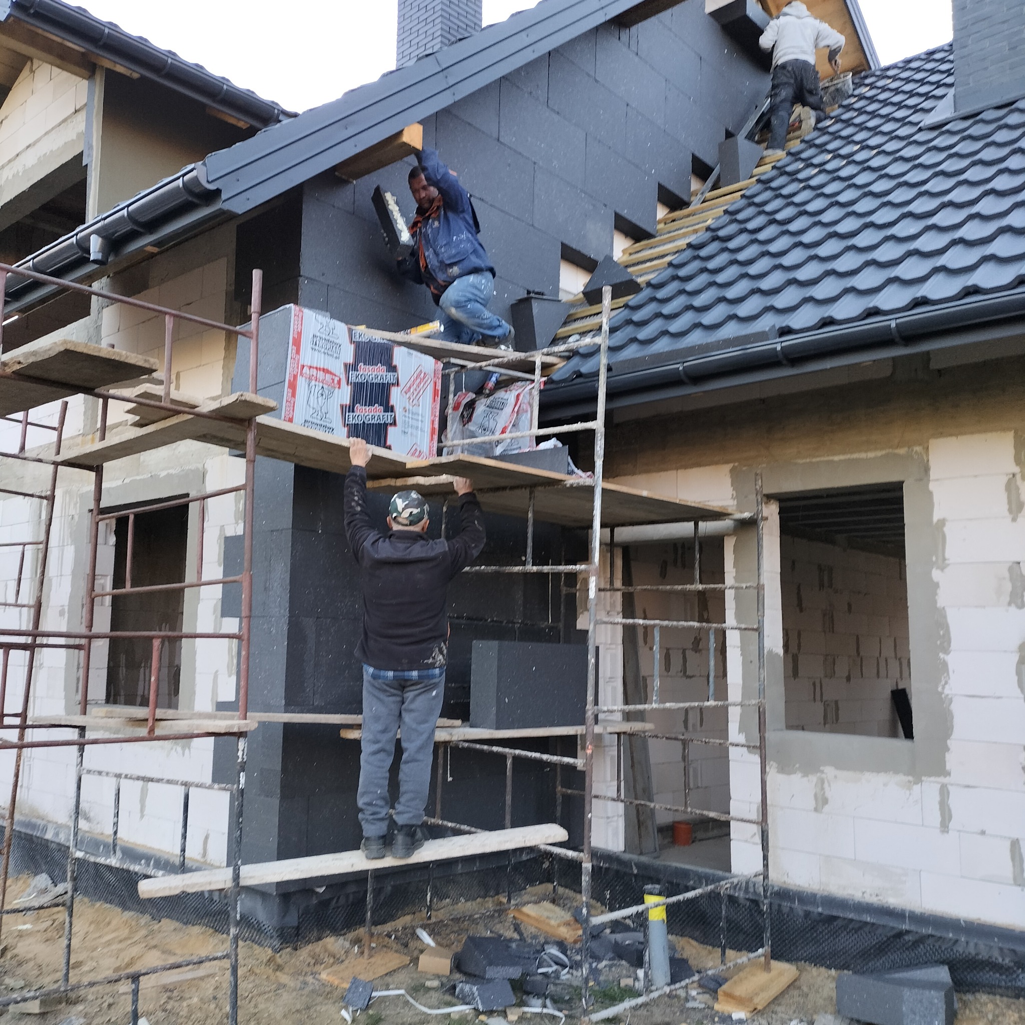 Prace ociepleniowe na elewacji domu jednorodzinnego. Robotnicy na rusztowaniu montują czarne płyty izolacyjne. Widoczne materiały izolacyjne i narzędzia.
