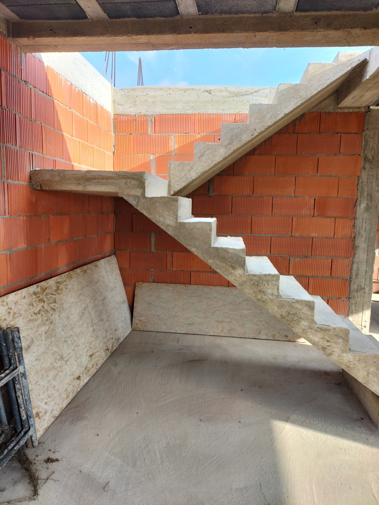 Surowe schody betonowe w trakcie budowy, widoczne ceglane ściany i fragment nieba przez otwór. Deski leżą na podłodze, a obok nich rusztowanie.