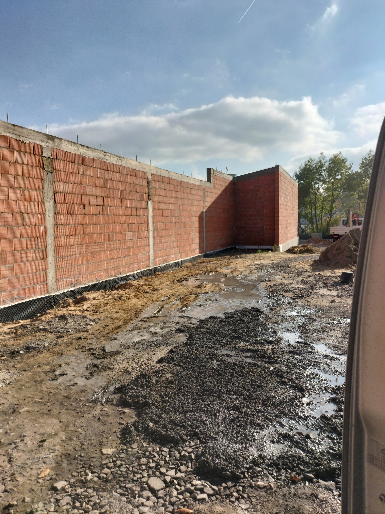 Wznoszenie muru z czerwonej cegły z widocznymi spoinami i izolacją przeciwwilgociową u podstawy, na tle nieba z chmurami i fragmentem pojazdu z prawej strony.