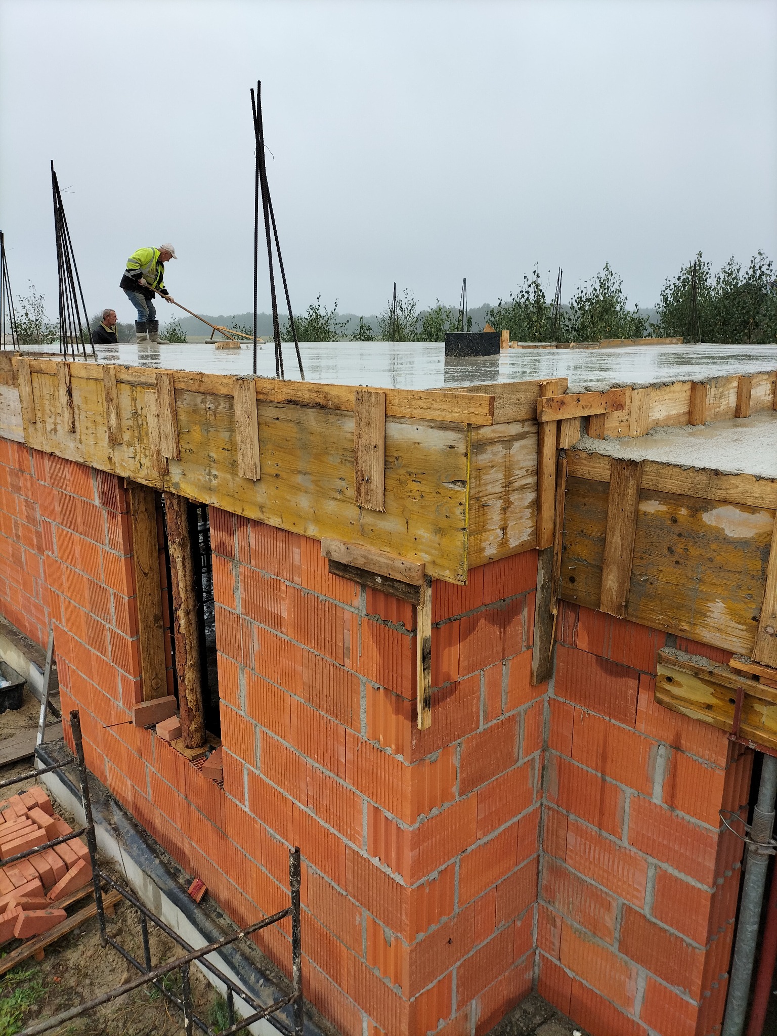 Murowanie ścian z czerwonej cegły, zalewanie betonem stropu z drewnianym szalunkiem. Dwóch pracowników w trakcie pracy na budowie w pochmurny dzień.