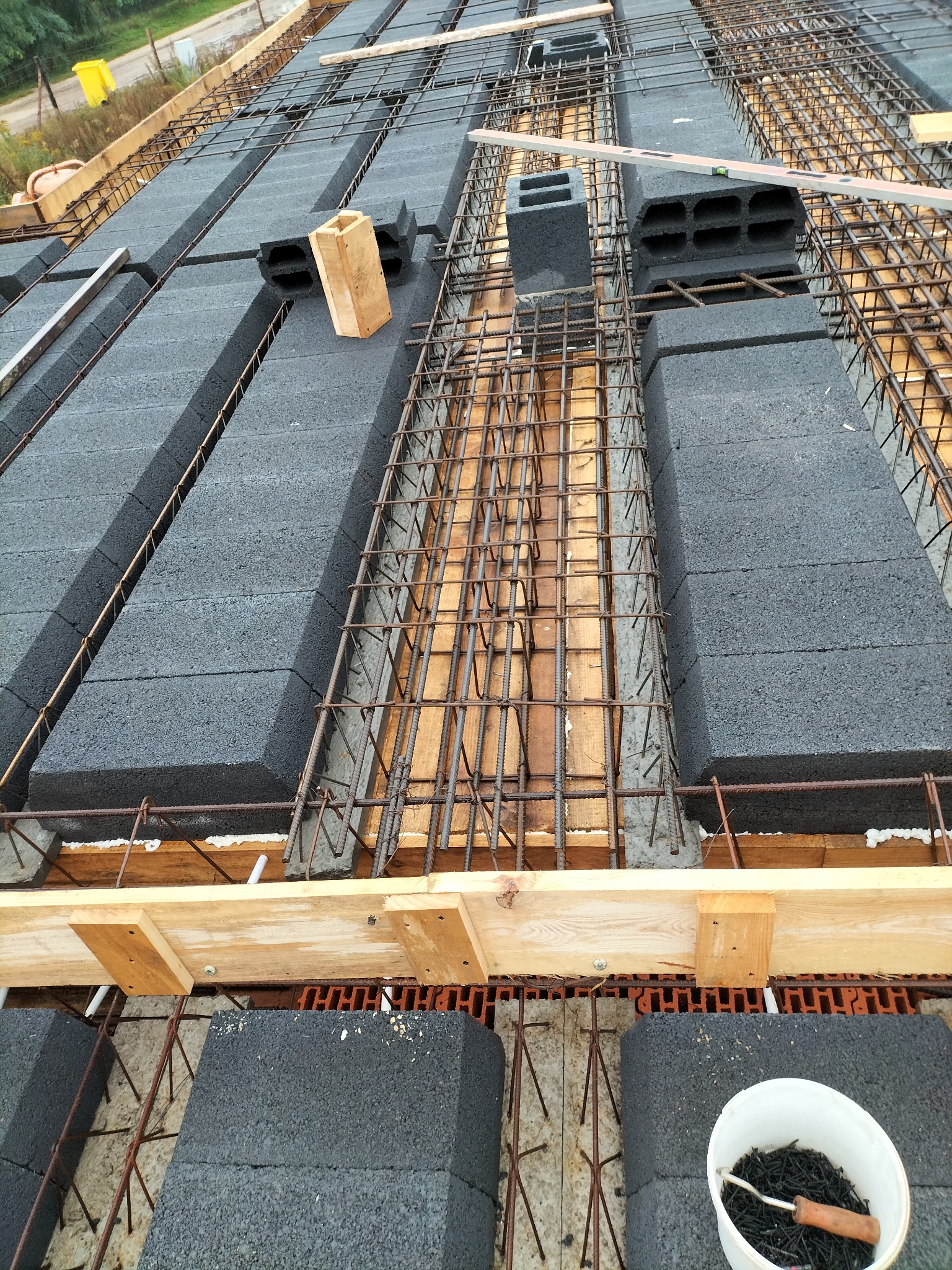 Budowa stropu: szare bloczki, zbrojenie z prętów, drewniane elementy szalunku. Widoczny poziomica i wiadro ze śrubami. Ujęcie z góry, w trakcie prac.