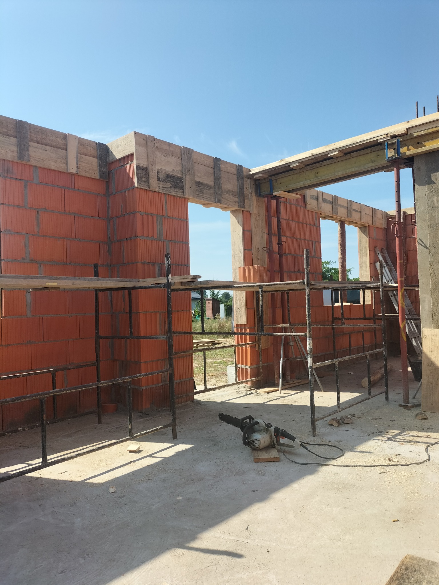 Wznoszenie ścian z czerwonej cegły ceramicznej, widoczne szalunki okienne i stropowe. Na pierwszym planie piła łańcuchowa i rusztowanie. Słoneczny dzień.