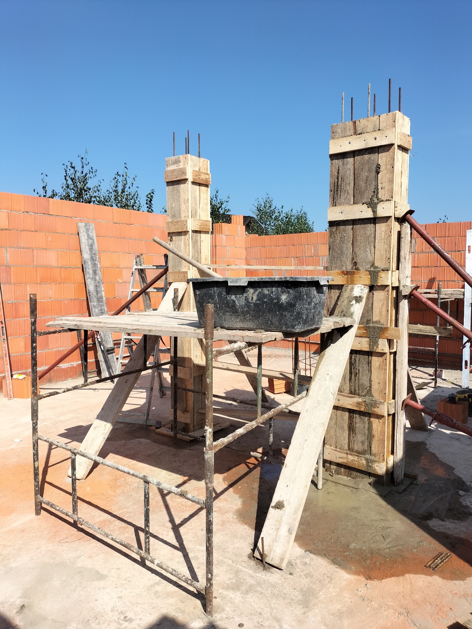 Szare rusztowanie, drewniane szalunki kolumn z wystającymi prętami zbrojeniowymi, czarna kuweta na zaprawę, mur z czerwonej cegły dziurawki w tle, błękitne niebo.