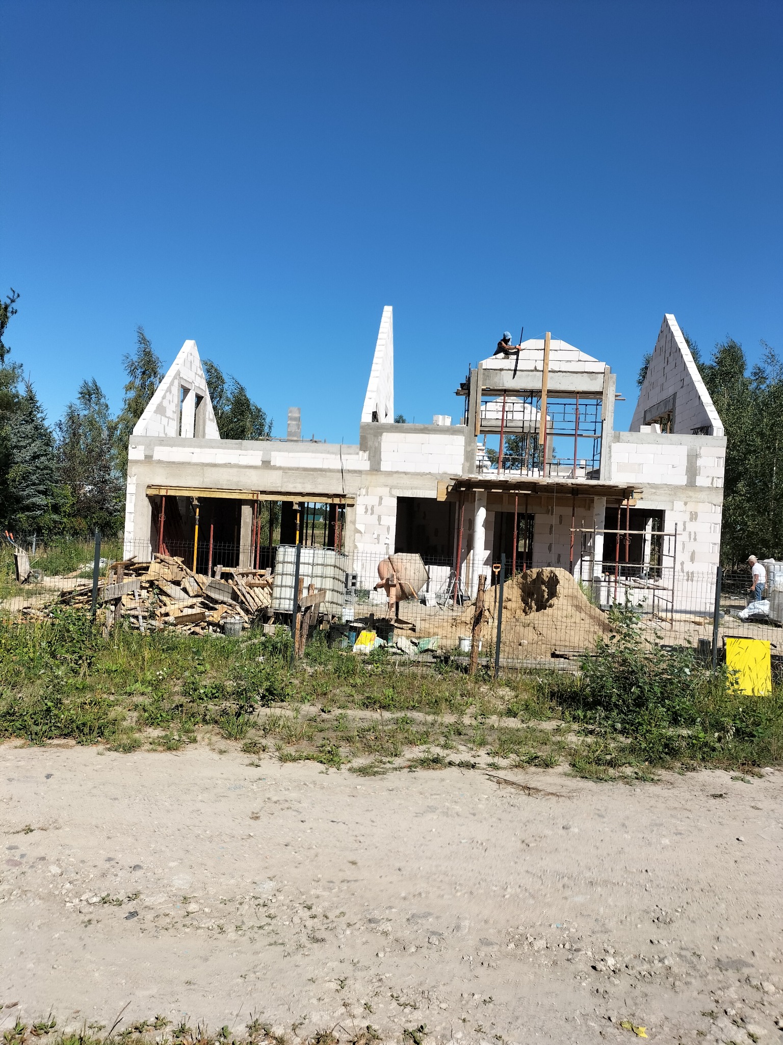 Budowa domu z białych bloczków, widoczny murarz na dachu, rusztowania, piasek i materiały budowlane. Niebieskie niebo w tle. Dom w stanie surowym otwartym.