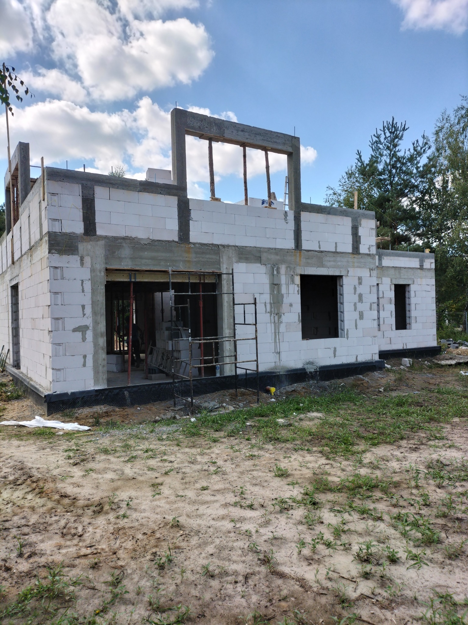 Niski, parterowy dom w trakcie budowy z białych bloczków, widoczne zbrojenia i szalunki okienne, na pierwszym planie piaszczyste podłoże i trawa.