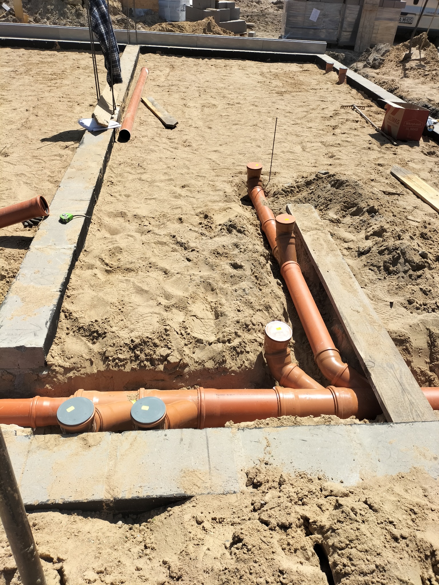 Instalacja kanalizacyjna w piasku, widoczne rury z szarymi pokrywami rewizyjnymi, częściowo zakopane w wykopie fundamentowym. Widok z góry, słoneczny dzień.