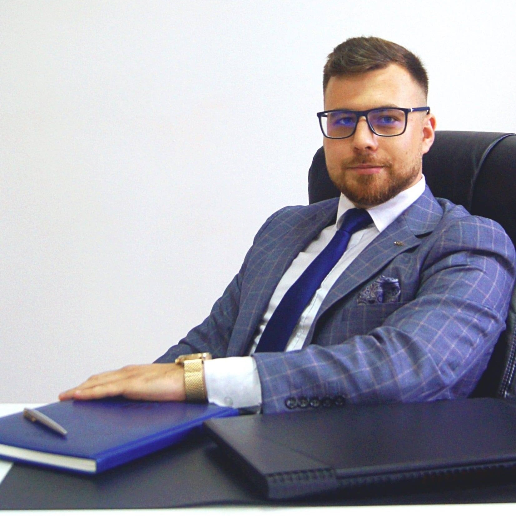 Portret mężczyzny w garniturze siedzącego za biurkiem z notatnikami i długopisem, patrzącego w obiektyw.