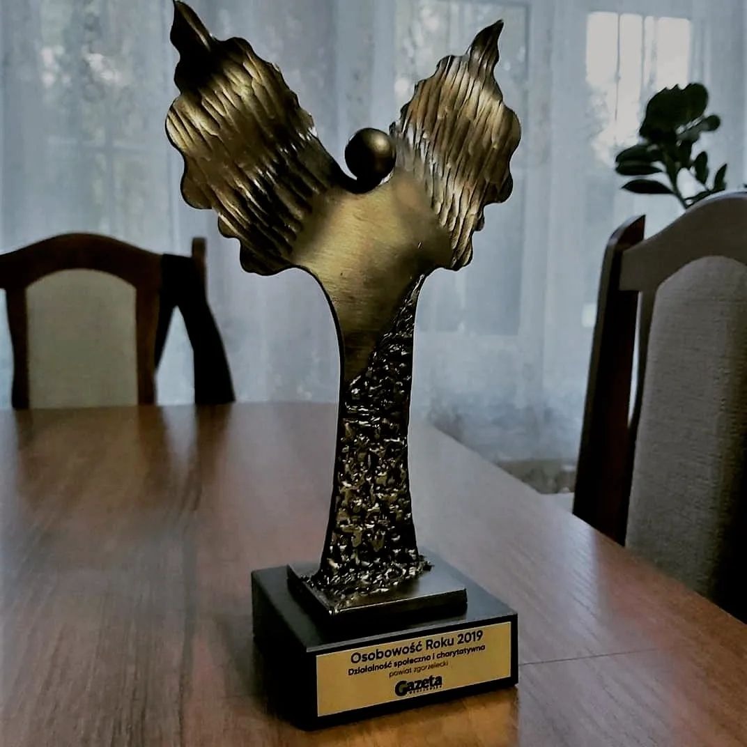 Statuetka 'Osobowość Roku 2019' z inskrypcją na drewnianym stole, widok z boku, w tle okno z zasłonami i roślina doniczkowa.