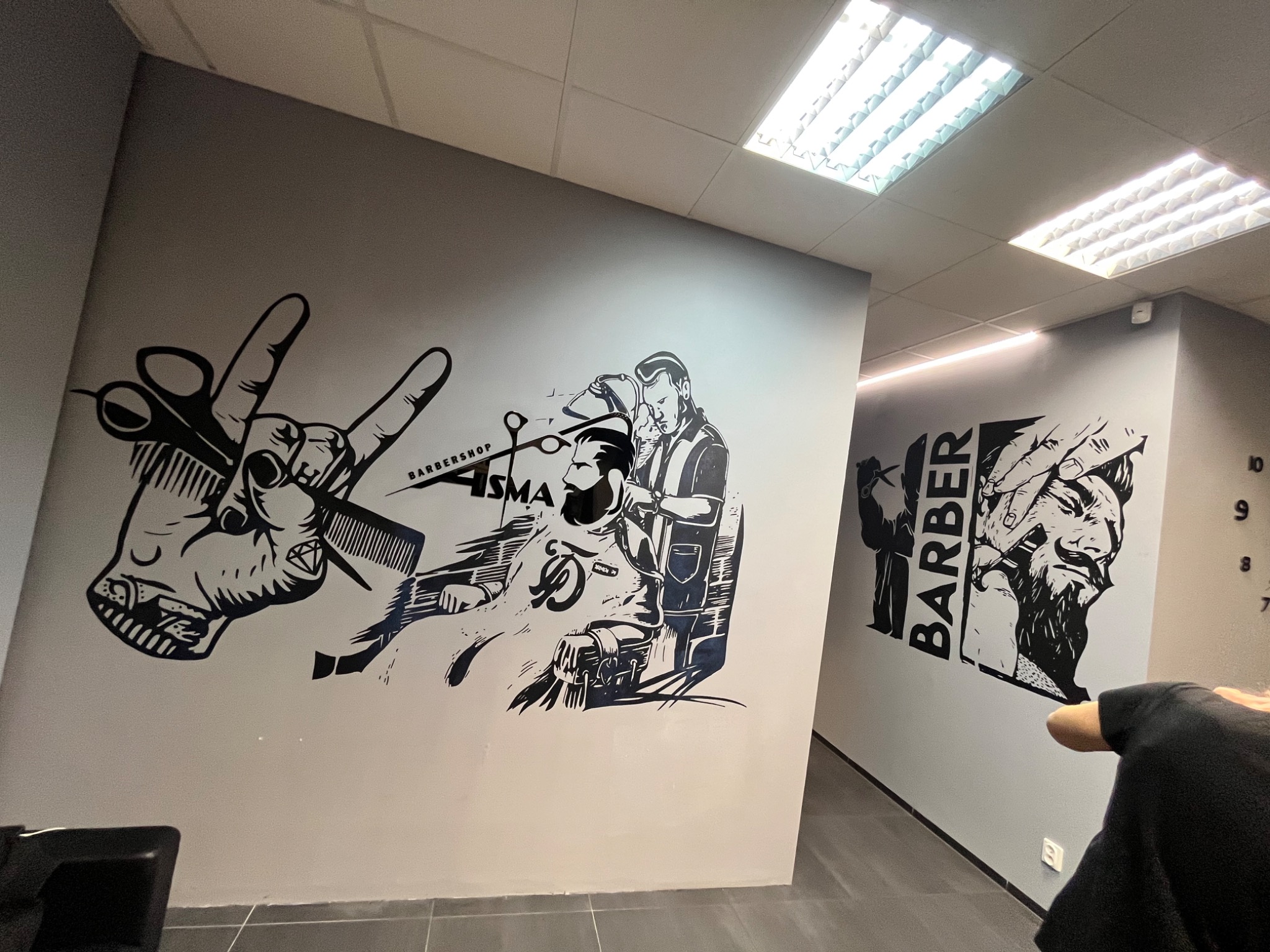 Ściana w salonie fryzjerskim ozdobiona czarno-białymi grafikami przedstawiającymi fryzjera i klientów, z elementami takimi jak nożyczki, grzebień i napis 'Barbershop'.
